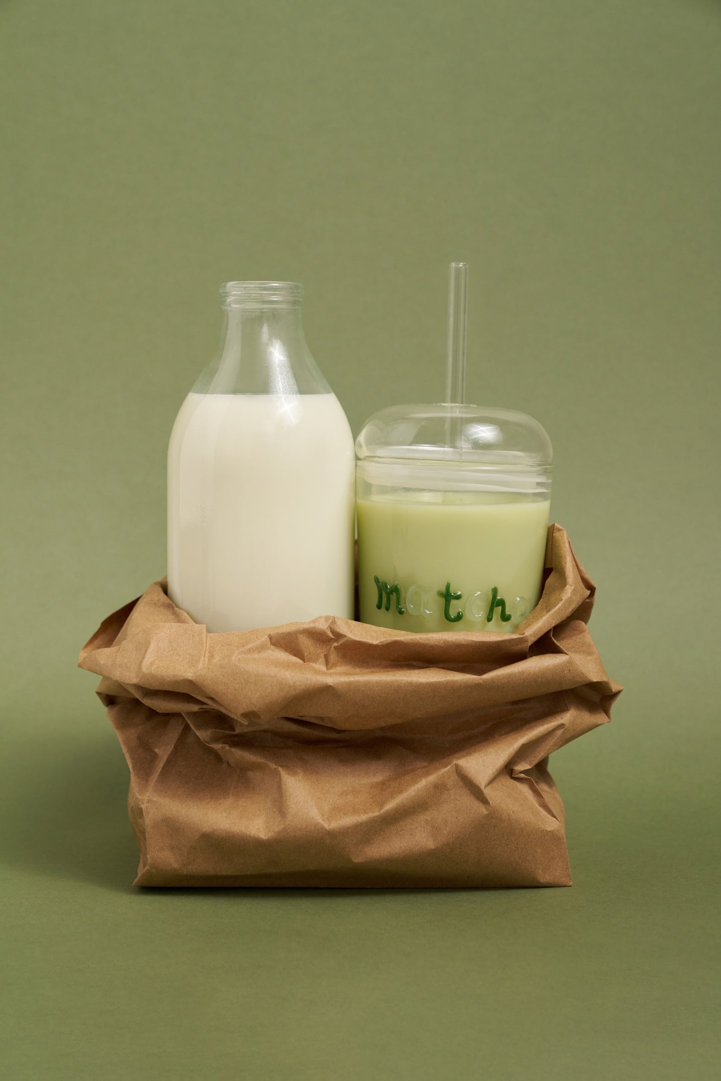 Milk bottle and glass with straw on a brown paper bag against a green background