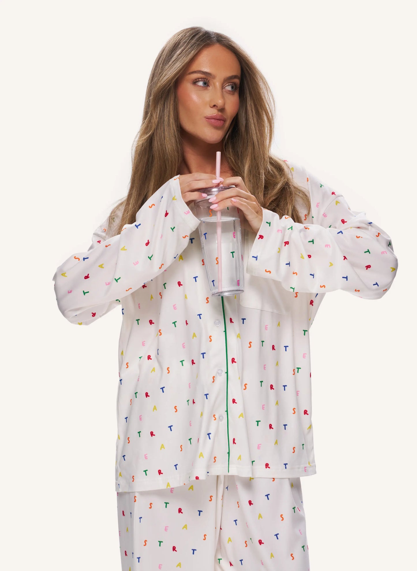 A woman wearing a white long-sleeve zip-up pajama top and matching pants, both covered in a small multicolor scattered print, holding a clear tumbler with a pink straw against a light beige studio background.