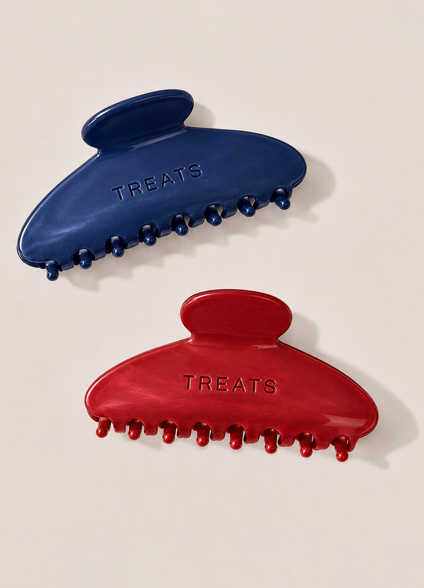 Two hair clips, one blue and one red, with 'TREATS' engraved on a beige background.