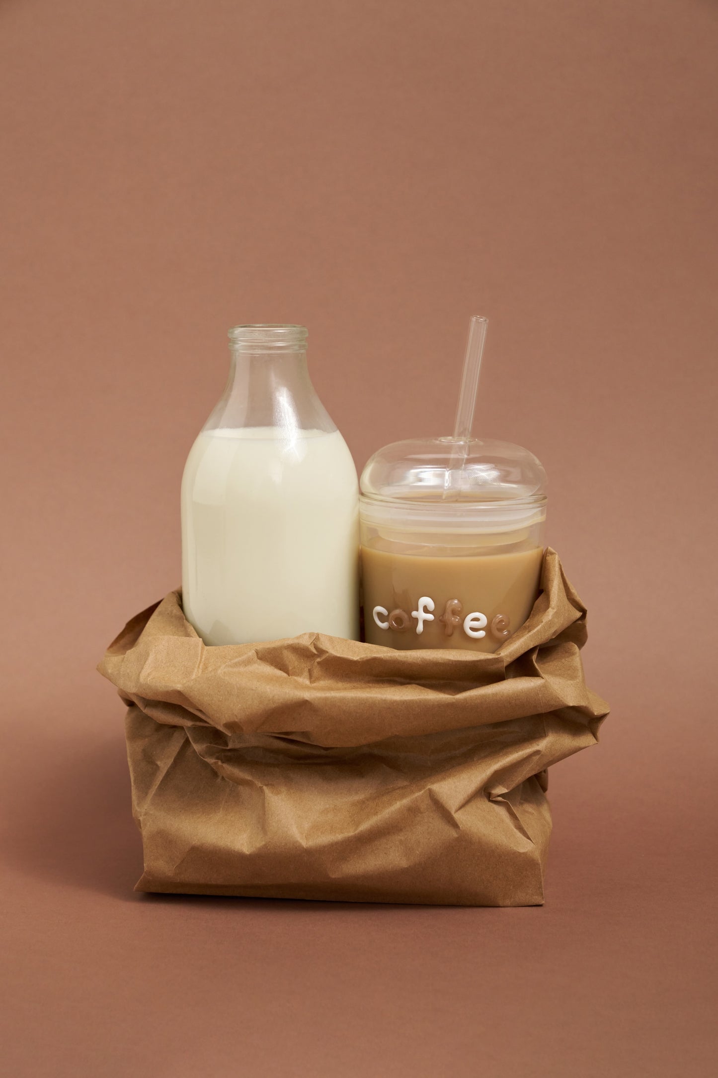 Glass bottle of milk and a cup of coffee with a straw on a brown paper bag against a brown background