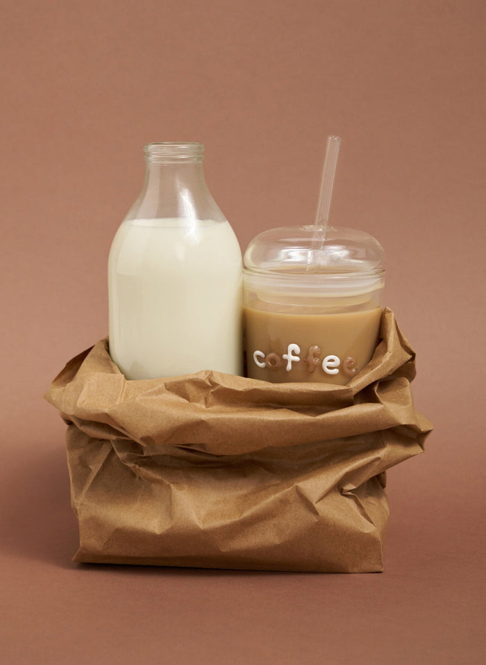 Glass bottle of milk and a cup of coffee with a straw on a brown paper bag against a brown background