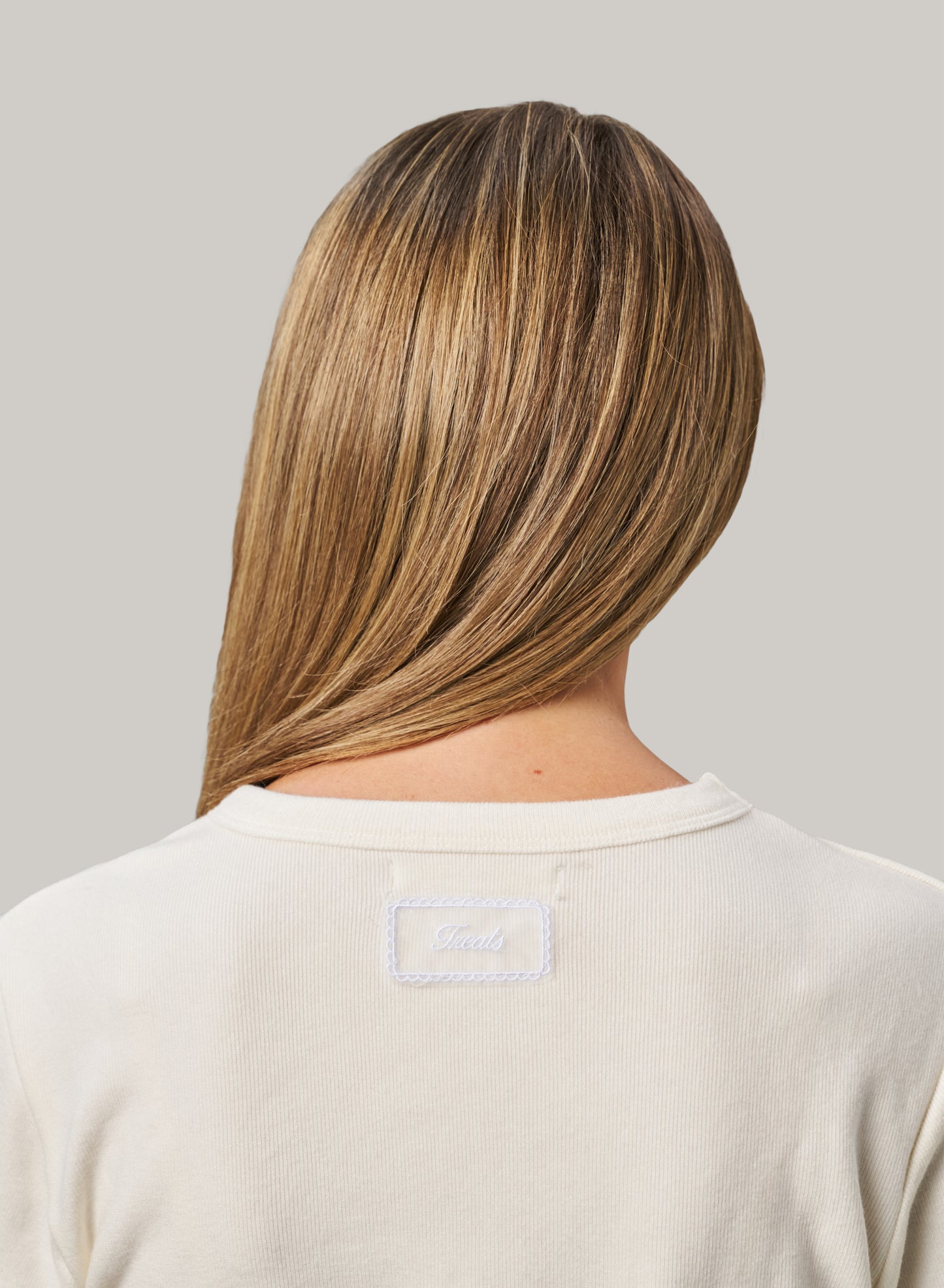 Person with long, straight hair wearing a beige shirt with a brand logo on a grey background
