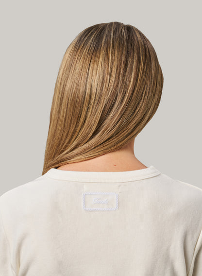 Person with long, straight hair wearing a beige shirt with a brand logo on a grey background