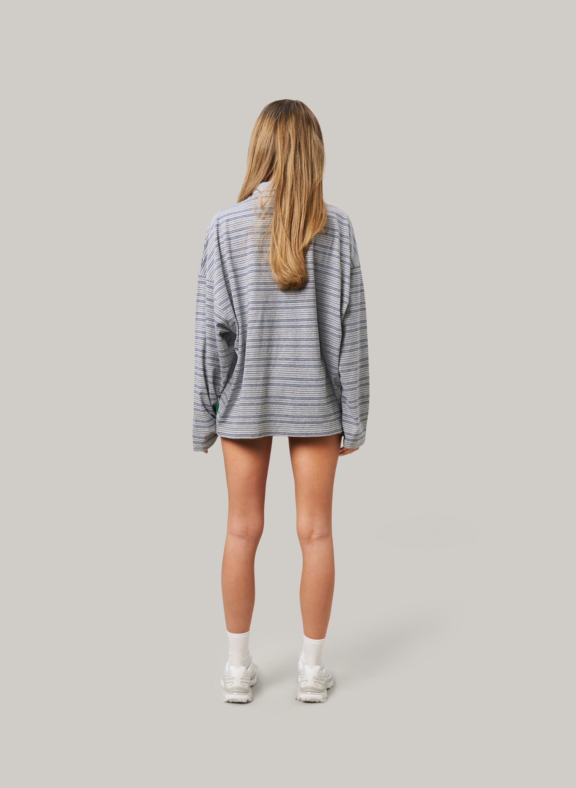 Woman facing backwards wearing an oversized grey striped long-sleeve shirt and shorts with white sneakers, standing against a plain background