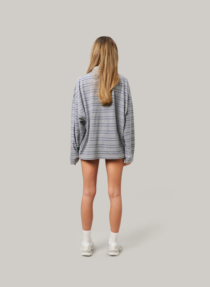 Woman facing backwards wearing an oversized grey striped long-sleeve shirt and shorts with white sneakers, standing against a plain background