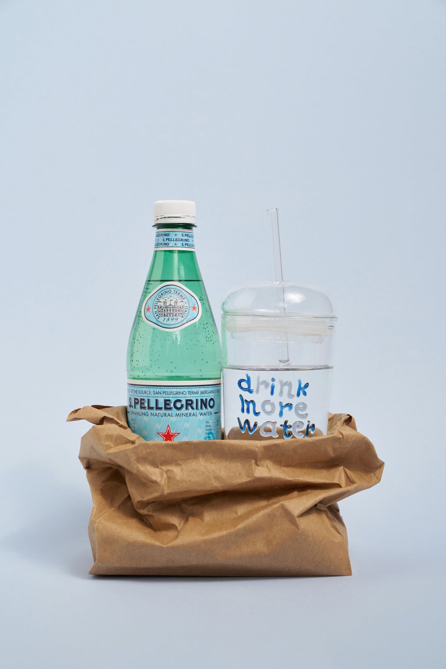 San Pellegrino bottle and 'drink more water' tumbler in a brown paper bag on a light blue background