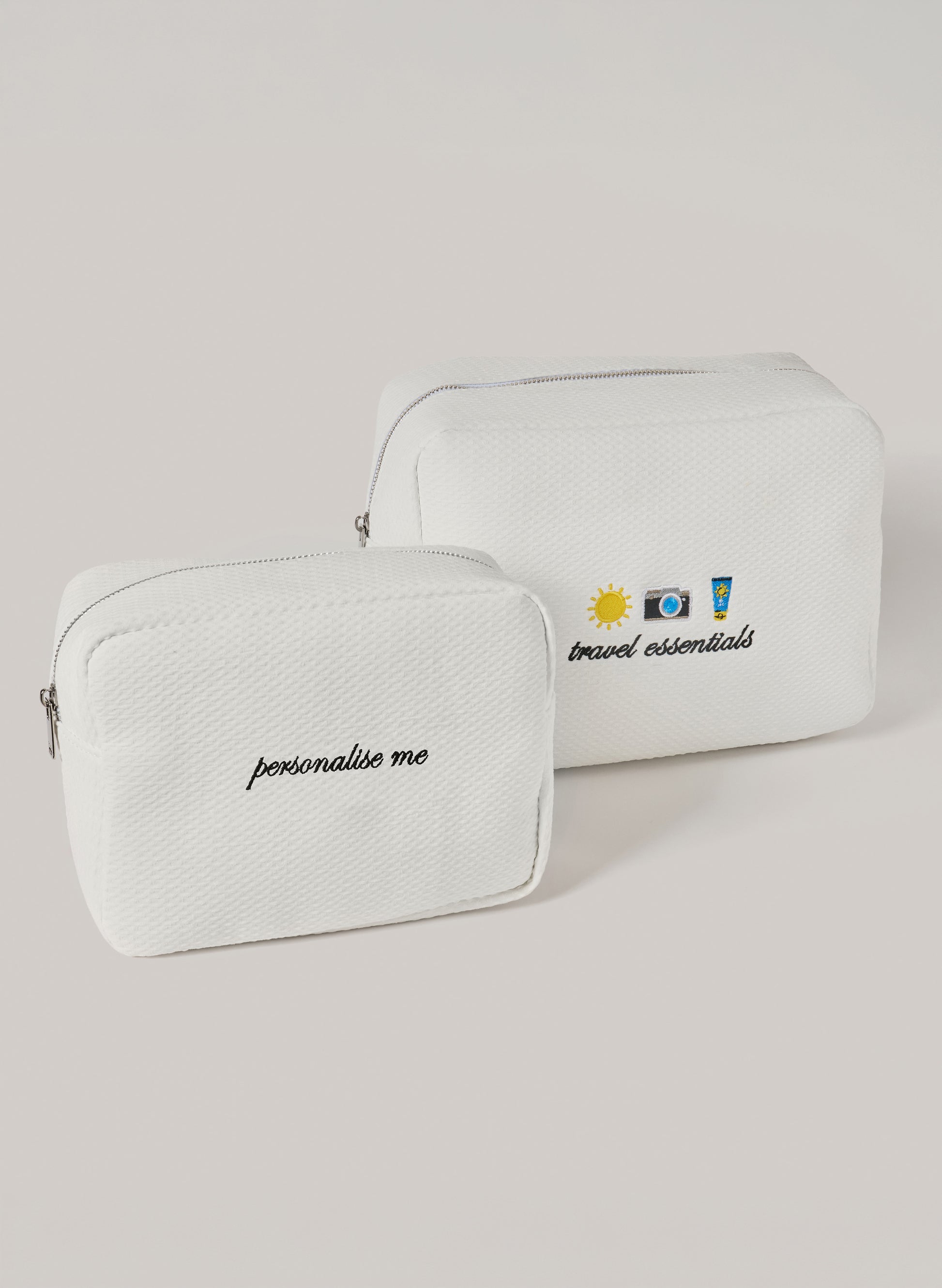 Two white waffle-textured pouches with zipper closures, one featuring ‘personalise me’ text and the other with embroidered travel icons and ‘travel essentials’ text on the front