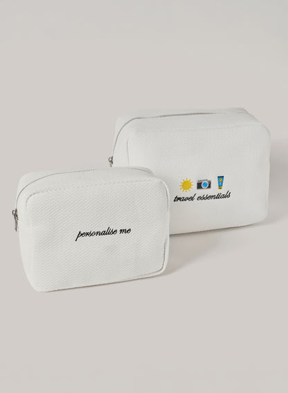 Two white waffle-textured pouches with zipper closures, one featuring ‘personalise me’ text and the other with embroidered travel icons and ‘travel essentials’ text on the front