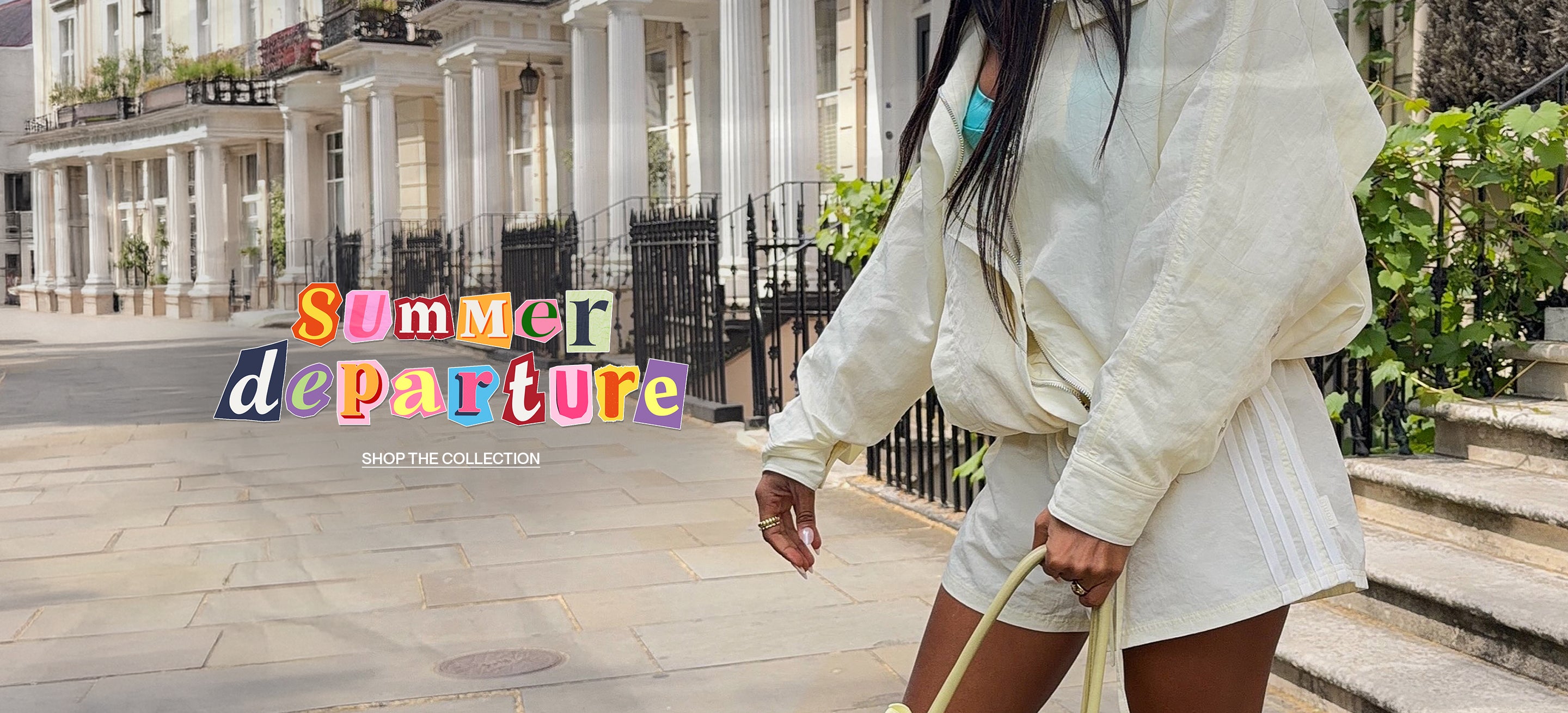 A woman wearing a cream-colored oversized long-sleeve top and matching shorts walks down a sunny, upscale street lined with classic white buildings. She is holding a light beige tote bag.
