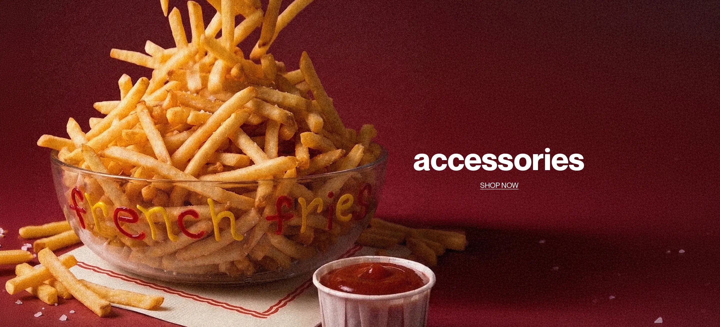 A clear glass bowl filled with french fries on a dark red background, with a small cup of ketchup in front and the word 'accessories' printed on the right side.