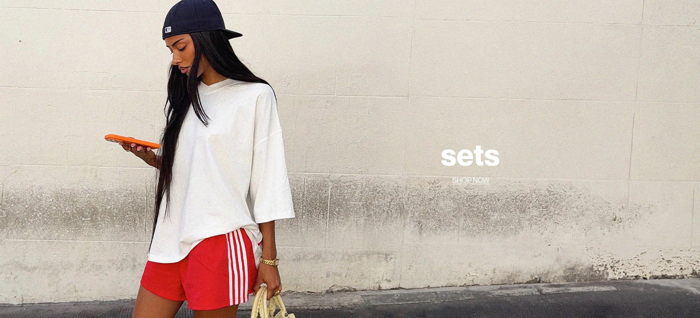A woman stands against a light concrete wall wearing a white oversized T-shirt, red athletic shorts with white stripes, a dark navy cap, and holding an orange phone and a beige handbag. The word 'sets' appears on the right side.