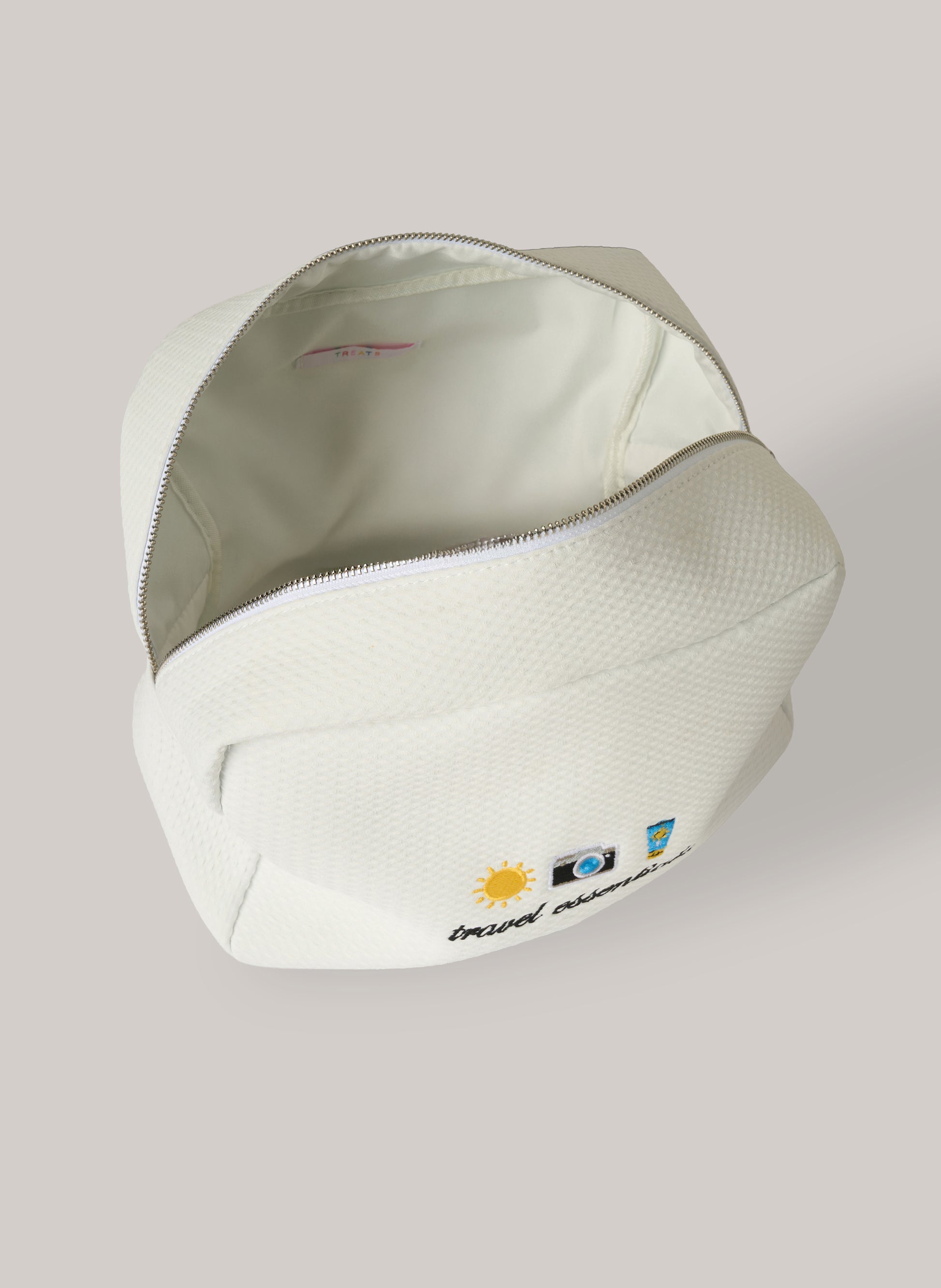 White waffle-textured pouch with zipper closure shown open, featuring small embroidered travel icons and the words ‘travel essentials’ on the front.