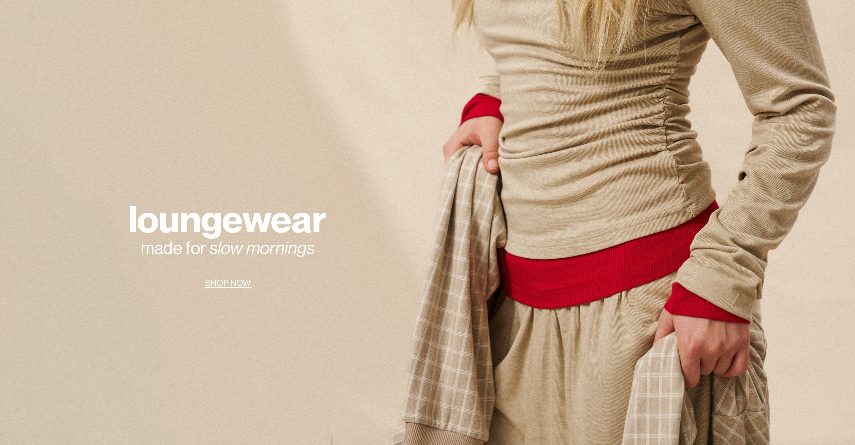 Person wearing beige loungewear with red accents, holding a plaid jacket on a beige background.