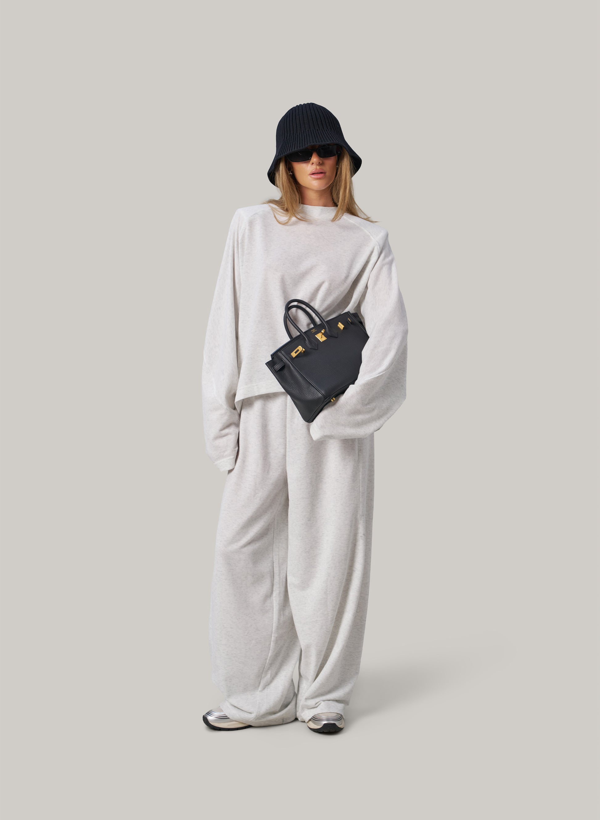 Person wearing a white outfit with a black hat and bag on a light grey background