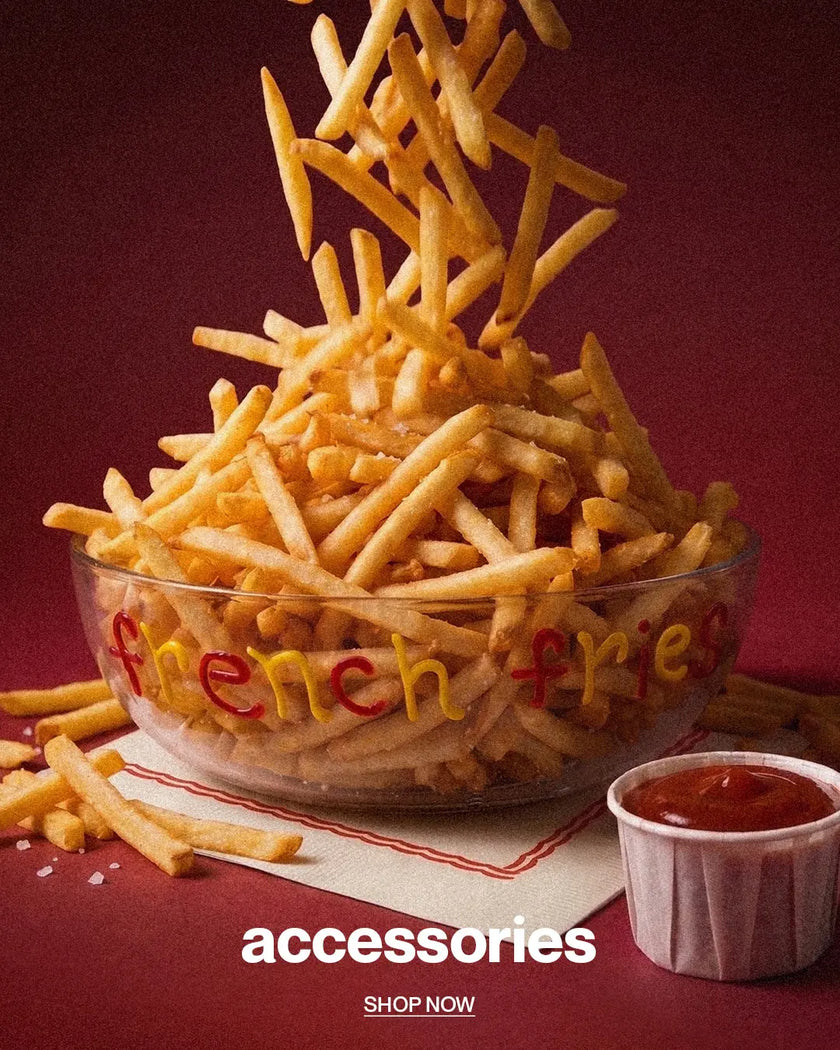 A clear glass bowl filled with golden french fries, with additional fries falling into the bowl against a dark burgundy background. The bowl has 'French fries' written on it in red and yellow letters.