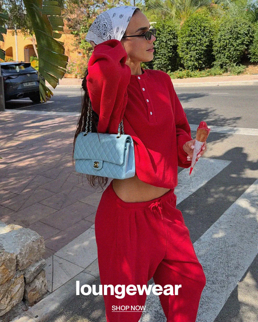 A woman wearing a matching red lounge set consisting of a cropped long-sleeve henley top and drawstring sweatpants, walking outdoors on a crosswalk. She is carrying a light blue quilted handbag and wearing a white patterned headscarf.