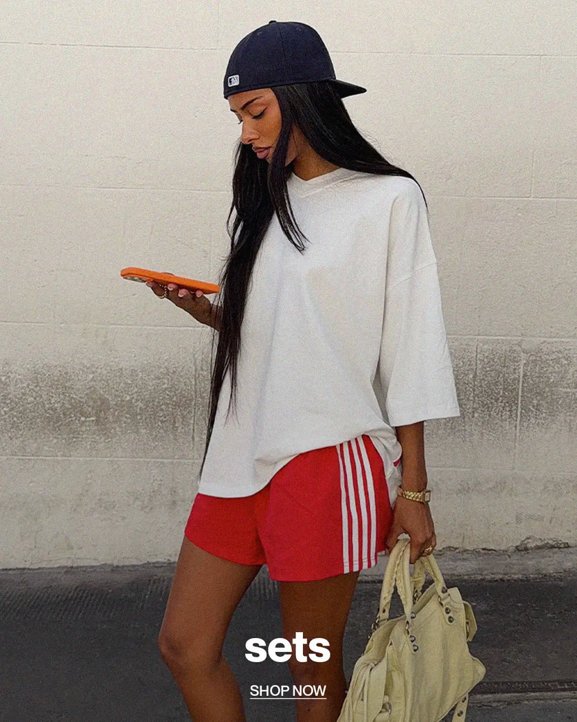 A woman wearing an oversized white t-shirt, red athletic shorts with white stripes, a navy baseball cap, and holding an orange phone and a beige handbag, standing against a concrete wall.