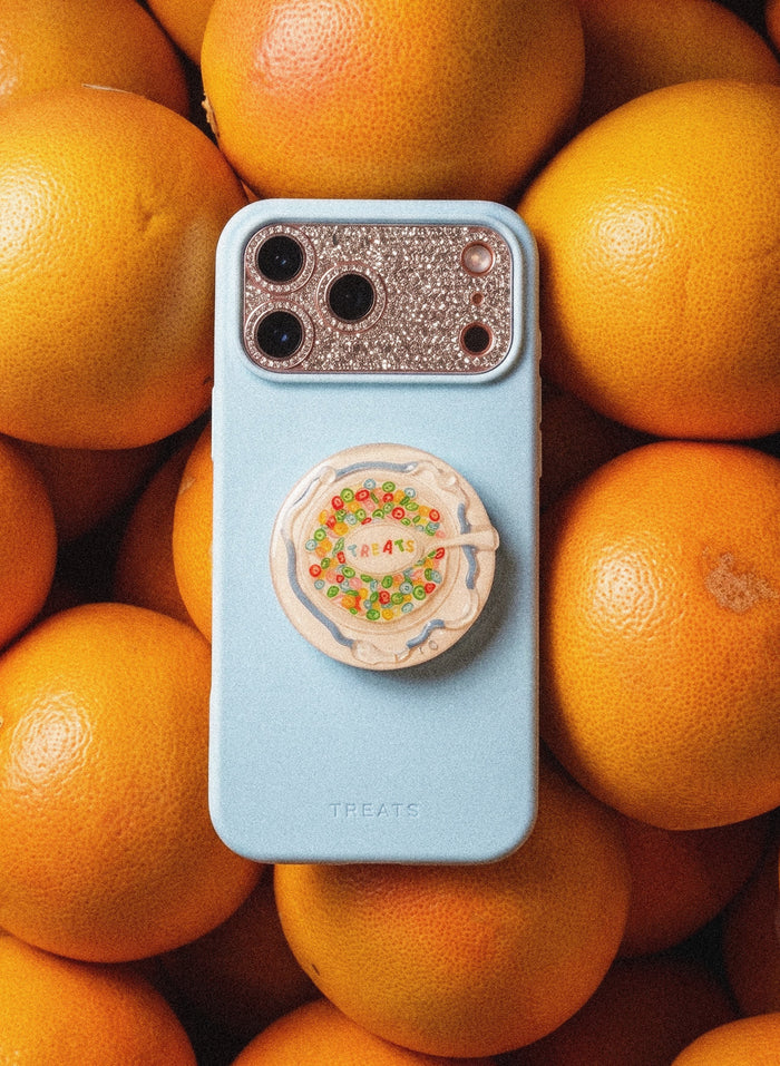 A light blue TREATS phone case with a glittery camera bezel and a round pop socket featuring a colorful floral design, centered on a background of fresh oranges.