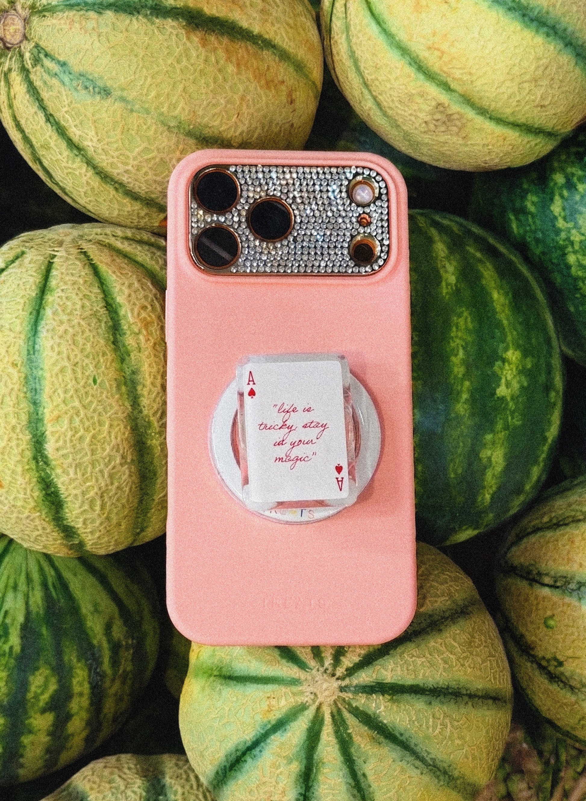 A pink phone case with a rhinestone-embellished camera cutout and a built-in white circular pop socket featuring a playing card design, shown resting on a bed of melons.