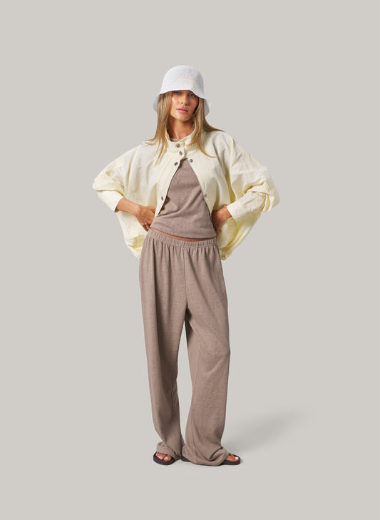Woman wearing a light-colored jacket, brown top, and pants on a plain background