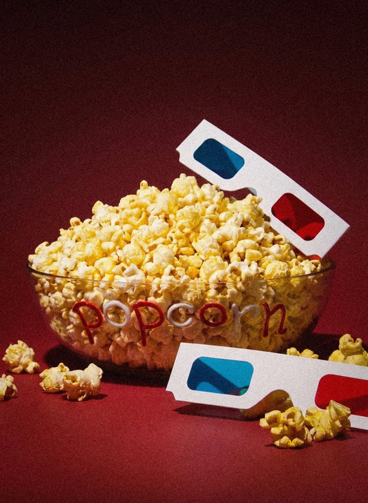 A clear glass bowl filled with popcorn on a dark red background, with two red-and-blue 3D glasses placed around it and a few scattered popcorn kernels.