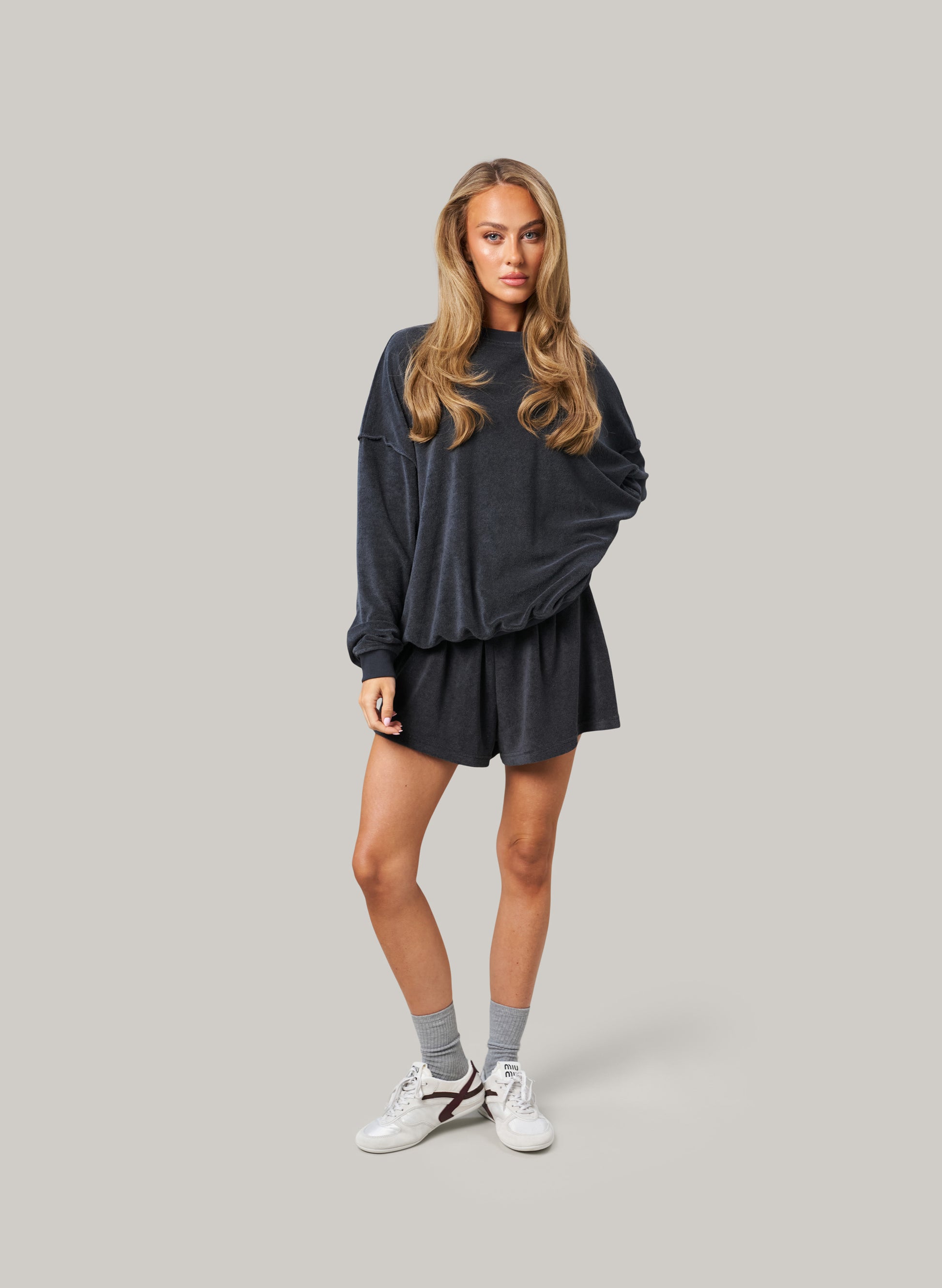 Woman wearing a dark oversized sweatshirt and shorts with white sneakers on a grey background