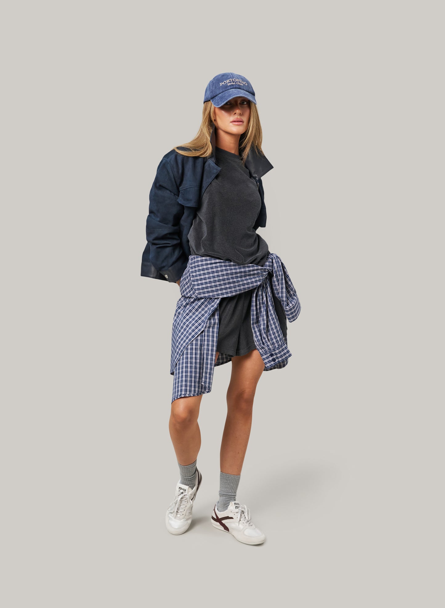 Person wearing a layered outfit with a jacket, shirt, skirt, and cap on a gray background