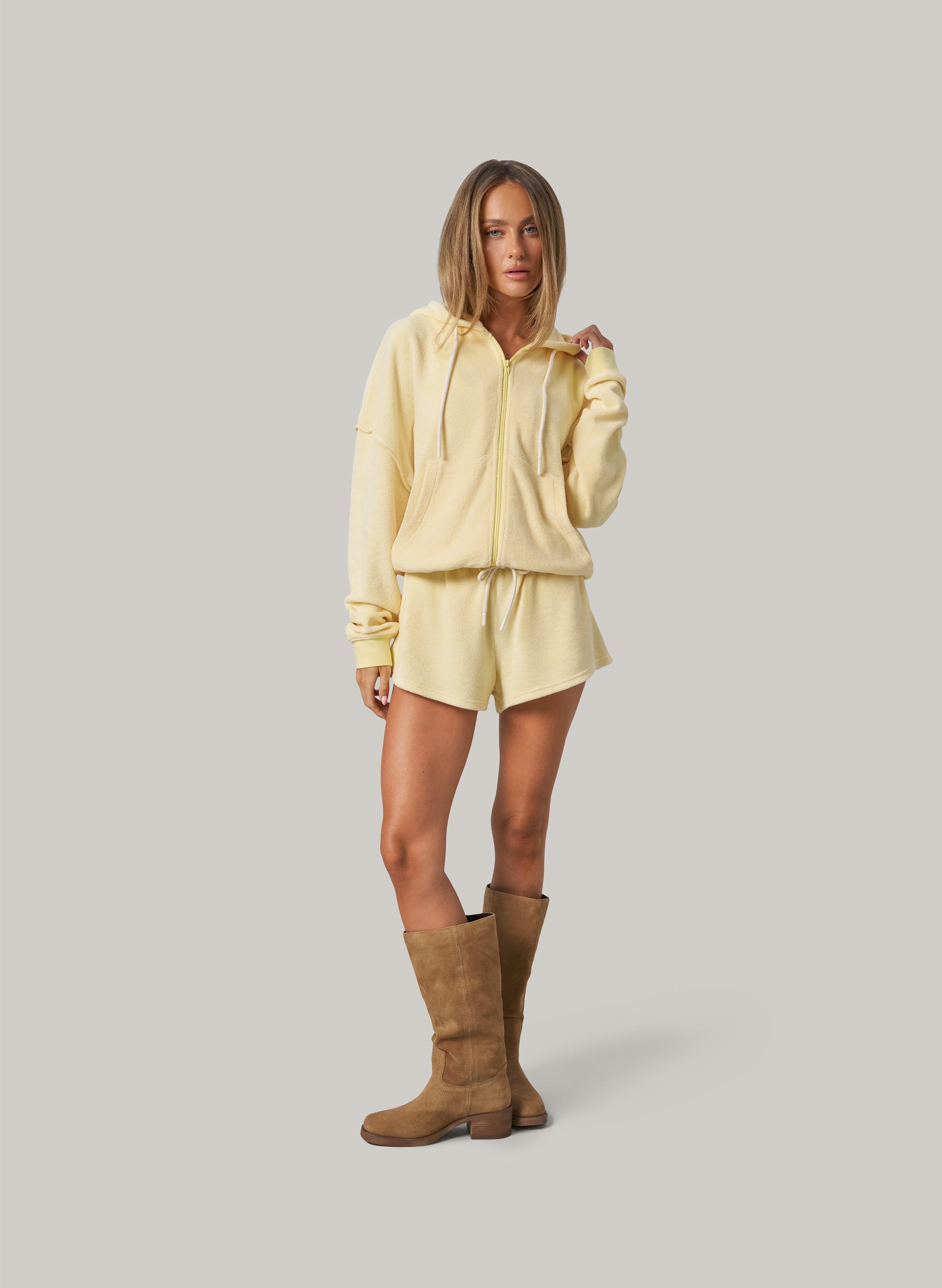 Person wearing a light yellow hoodie and shorts set with brown knee-high boots on a plain background