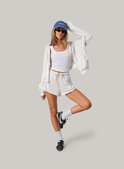 Woman in a white outfit with a blue cap and sunglasses on a light gray background