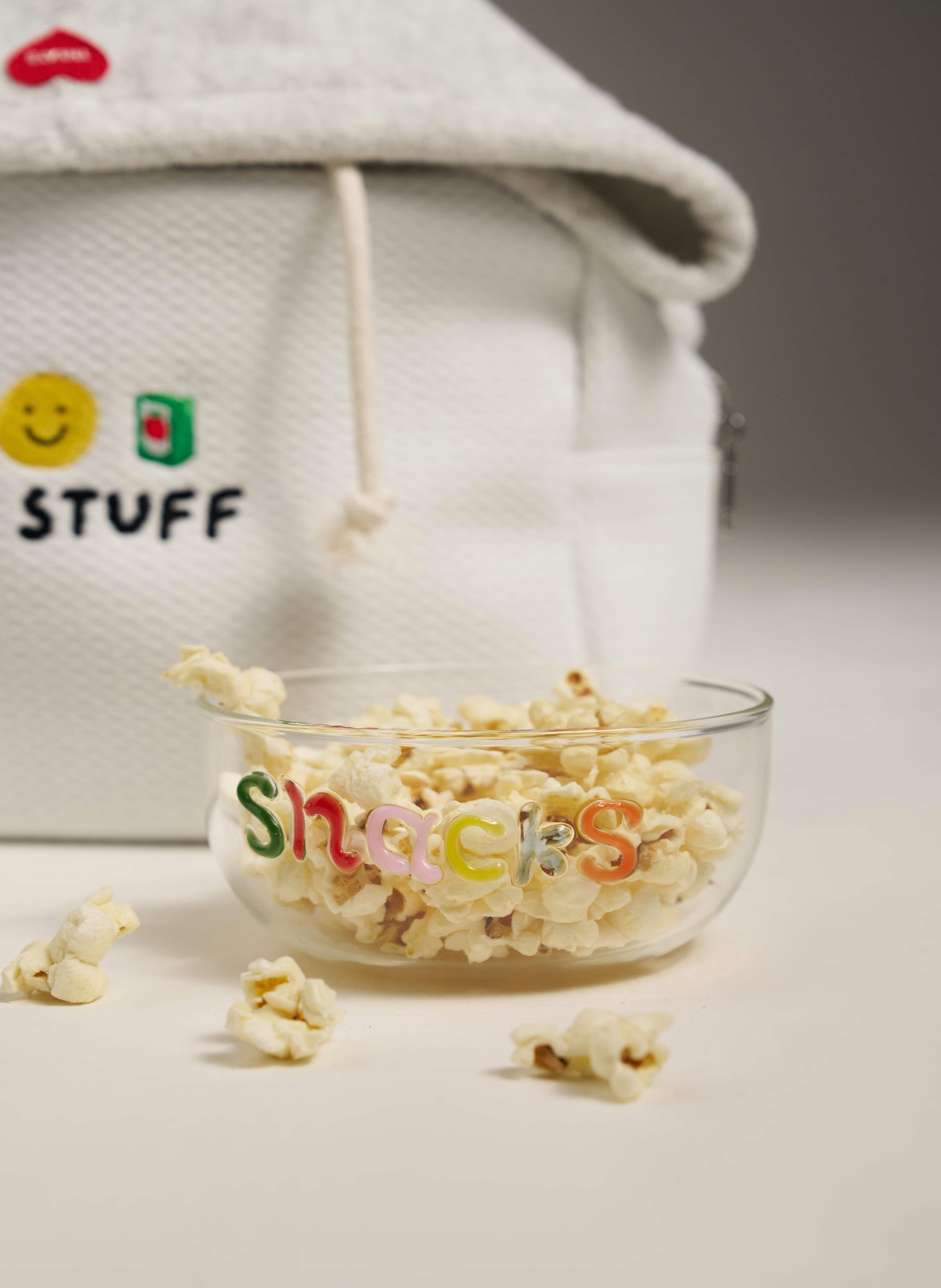 Glass bowl with popcorn labeled 'Snacks' in front of a white bag with colorful text.
