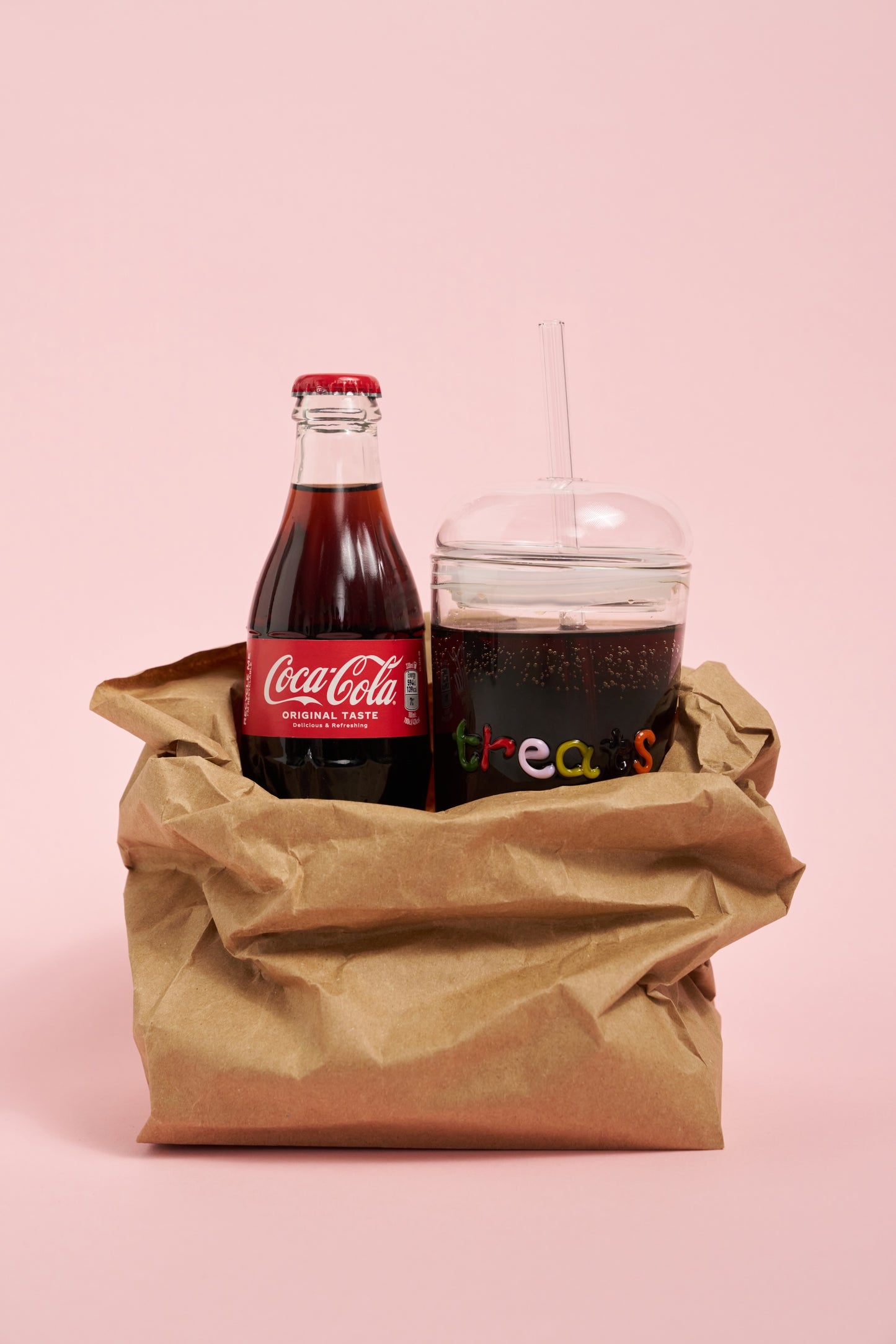 Coca-Cola bottle and a glass with a dark beverage in a brown paper bag on a pink background