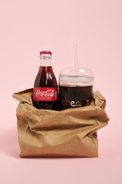 Coca-Cola bottle and a glass with a dark beverage in a brown paper bag on a pink background