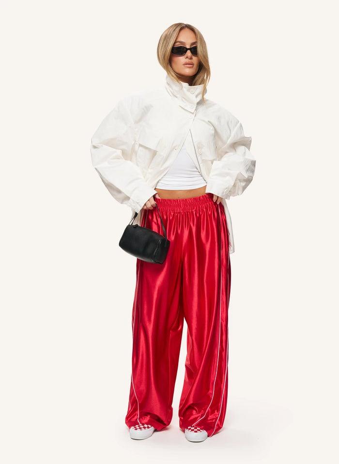 A model wearing a white cropped puffer jacket, a white ribbed top, shiny red wide-leg pants, white sneakers, and holding a small black handbag against a solid off-white background.