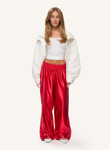 A model stands against a plain off-white studio background wearing shiny bright red wide-leg pants, a white cropped tank top, and an oversized white jacket.