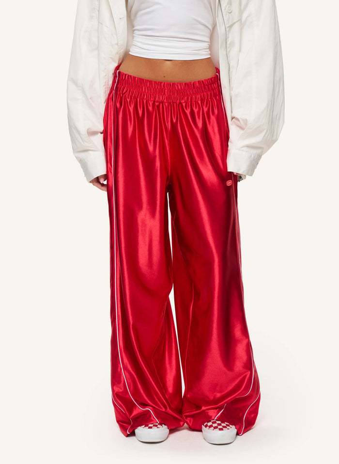 A model wearing shiny red wide-leg satin pants with a white cropped top and an open white jacket, shown from the waist down against a plain light background.