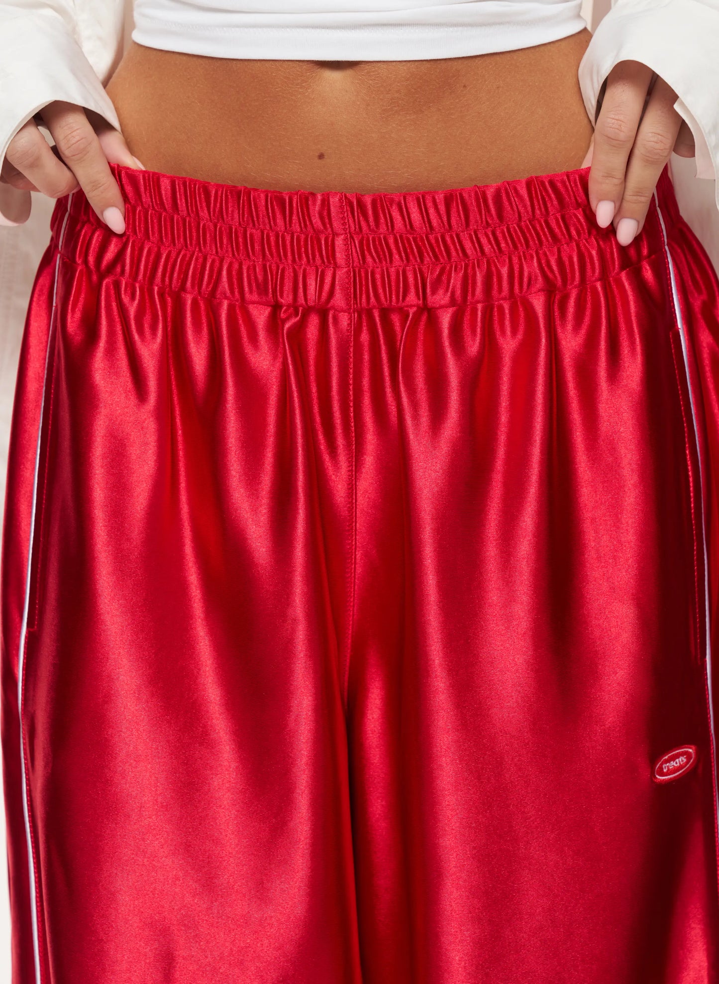 Close-up of a person wearing shiny red satin athletic pants with an elastic waistband and a small oval logo near the lower right leg. The person is also wearing a white cropped sweatshirt, with hands holding the waistband open.
