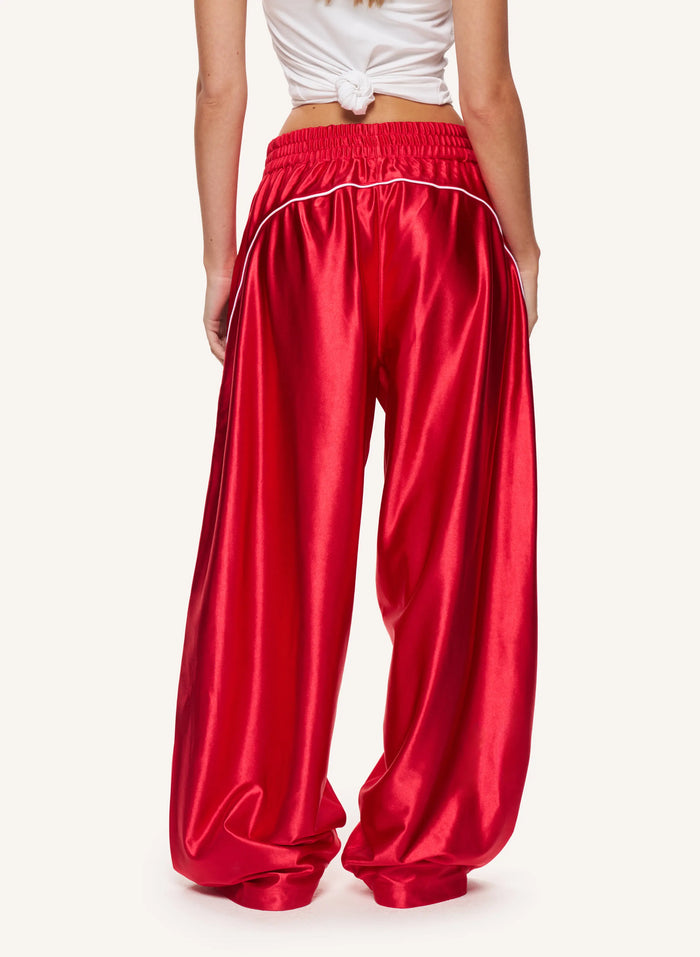 A model from the back wearing shiny red wide-leg pants with white piping along the waistband, paired with a white tied crop top, against a light off-white background.