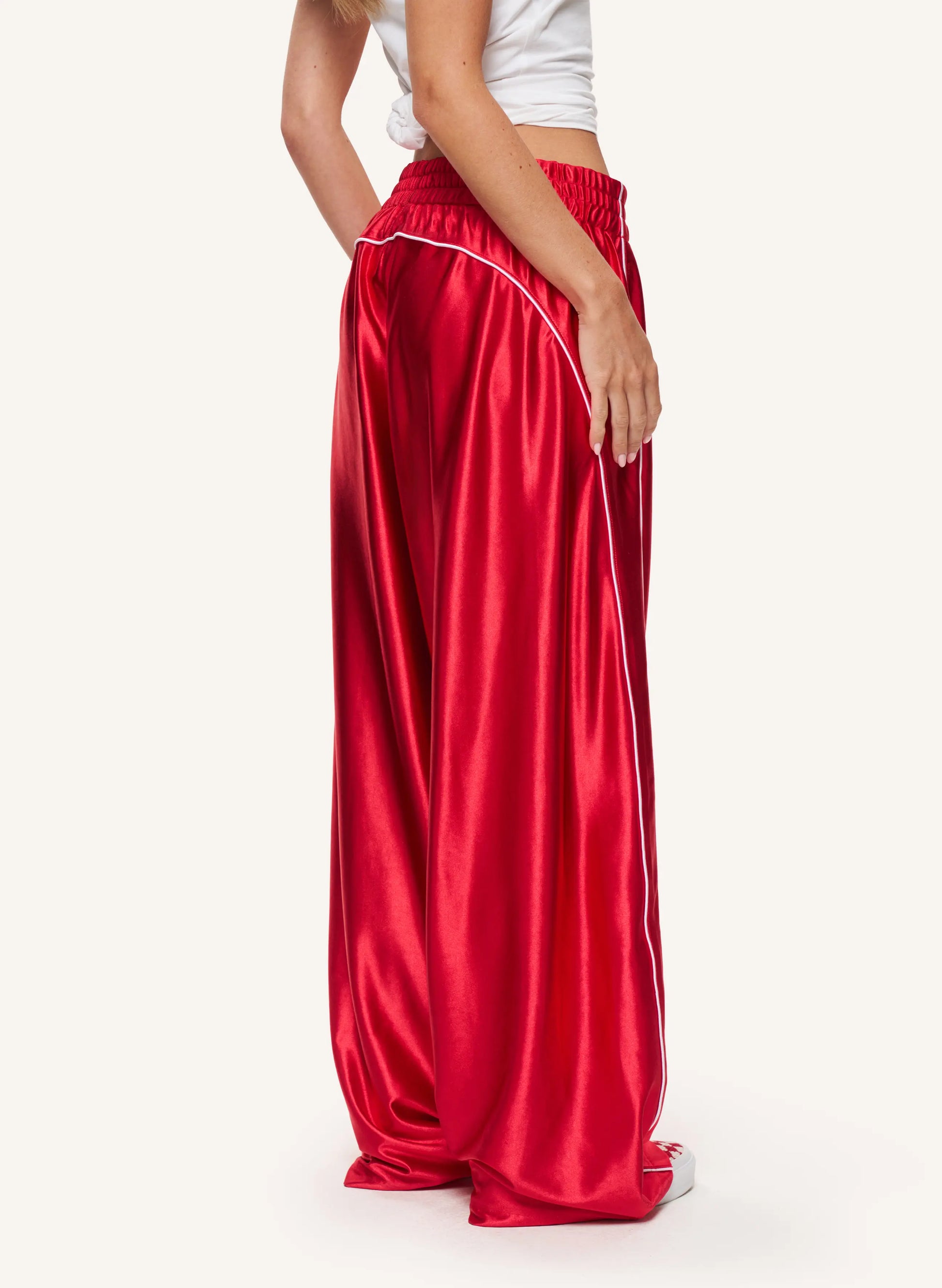 A model is shown from the back wearing shiny red wide-leg satin pants with a white side stripe, paired with a white crop top and white sneakers, against a plain off-white background.