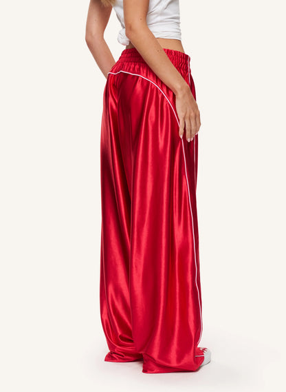 A model is shown from the back wearing shiny red wide-leg satin pants with a white side stripe, paired with a white crop top and white sneakers, against a plain off-white background.