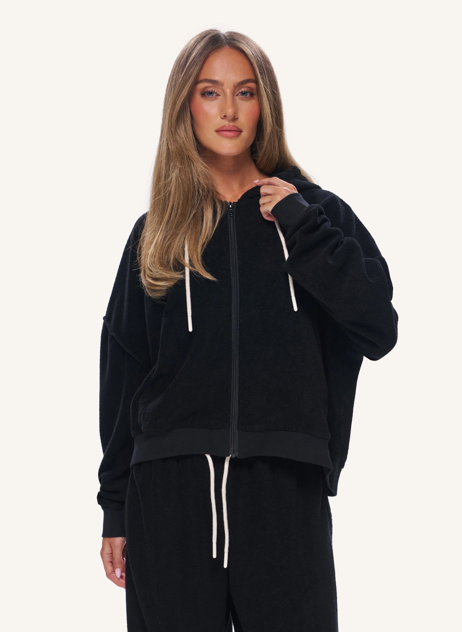 A woman modeling a black zip-up hoodie with white drawstrings, paired with matching black sweatpants, against a plain light background.