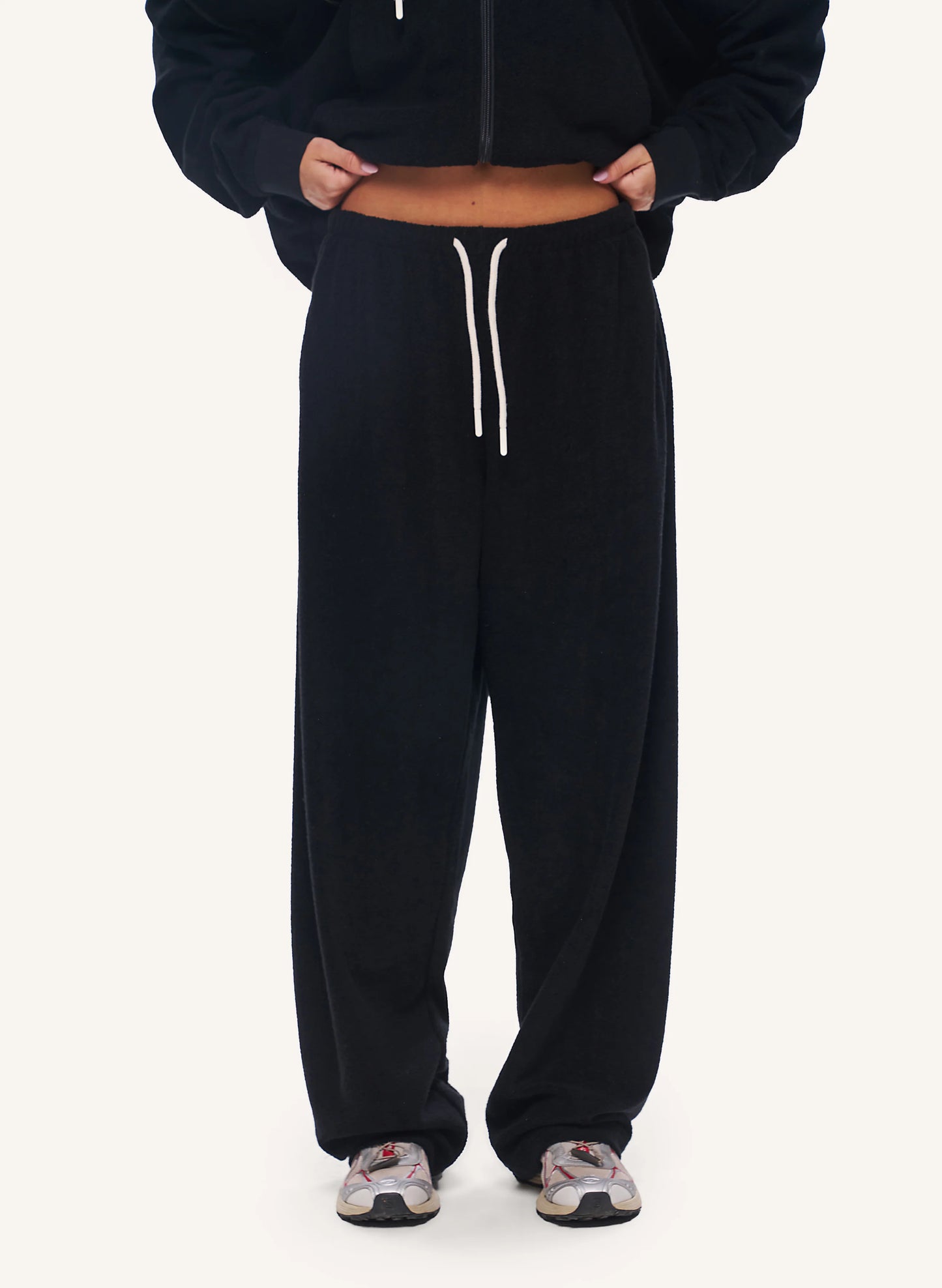 A person wearing black wide-leg sweatpants with a white drawstring, styled with a black cropped zip-up hoodie and patterned sneakers, shown from the chest down on a light off-white background.