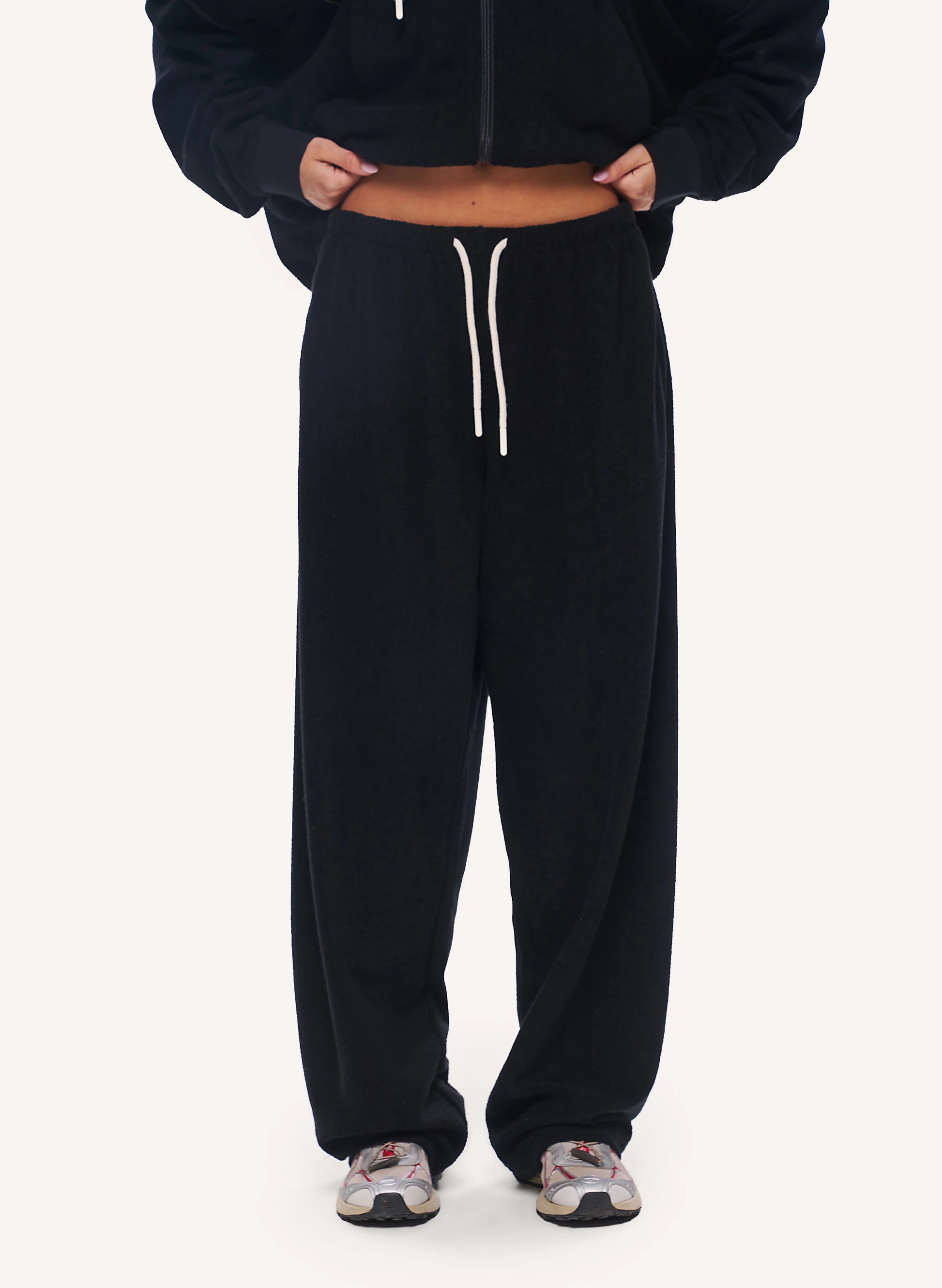 A person wearing black wide-leg sweatpants with a white drawstring, styled with a black cropped zip-up hoodie and patterned sneakers, shown from the chest down on a light off-white background.