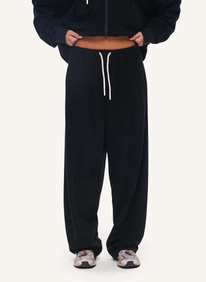 A person wearing black wide-leg sweatpants with a white drawstring, styled with a black cropped zip-up hoodie and patterned sneakers, shown from the chest down on a light off-white background.