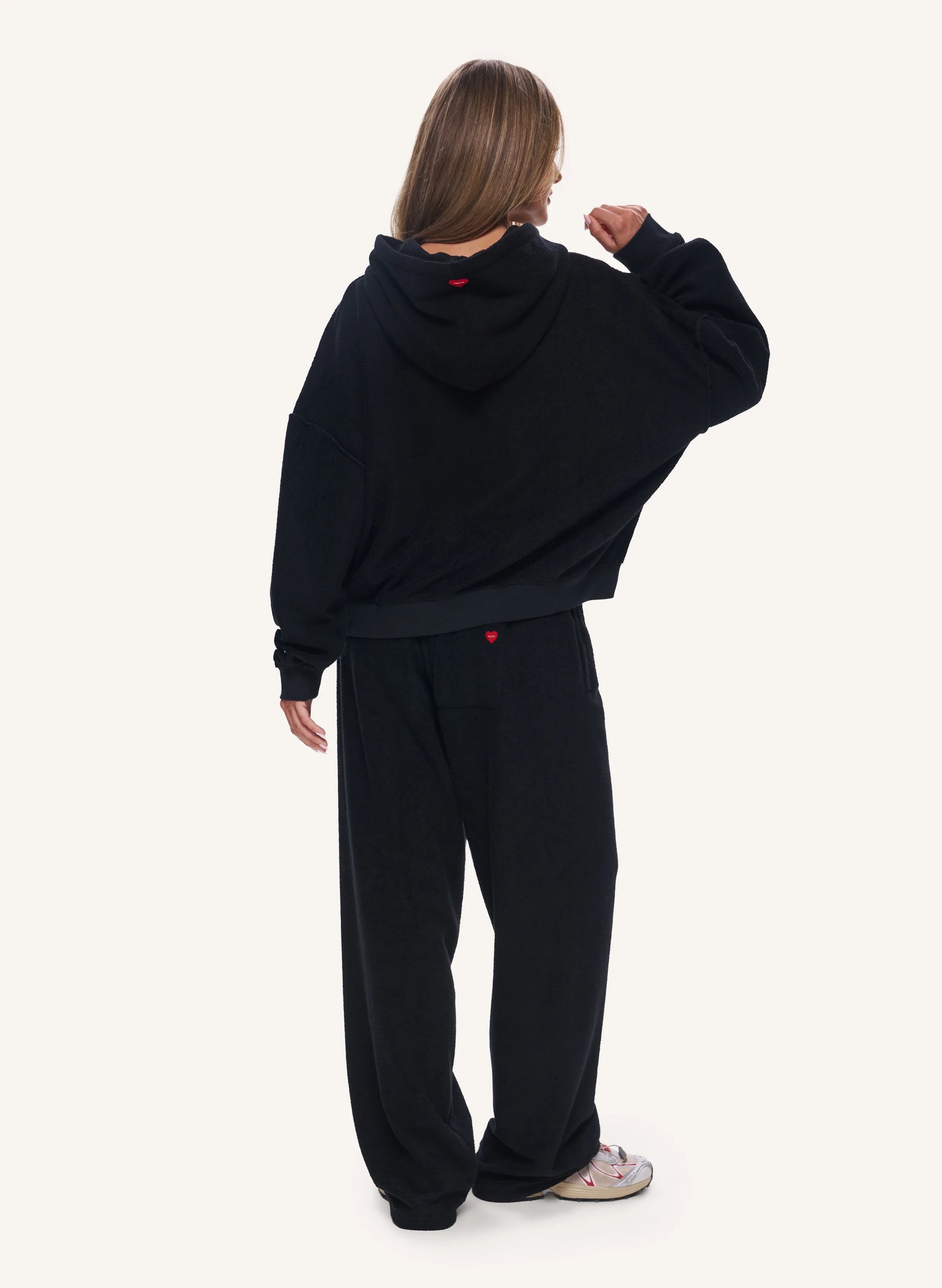 A model shown from the back wearing a black oversized cropped hoodie and matching black sweatpants, with a small red logo detail on the back of the hoodie and pants. The background is a solid off-white studio backdrop.