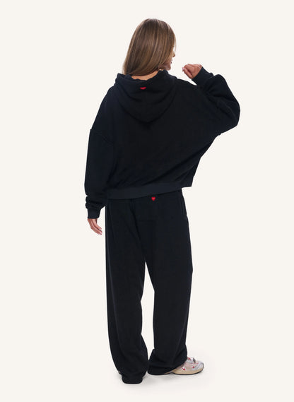 A model shown from the back wearing a black oversized cropped hoodie and matching black sweatpants, with a small red logo detail on the back of the hoodie and pants. The background is a solid off-white studio backdrop.