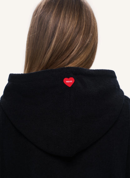 Back view of a person wearing a black hooded sweatshirt, showing a small red heart-shaped patch with the word 'TREATS' on the back of the neck.