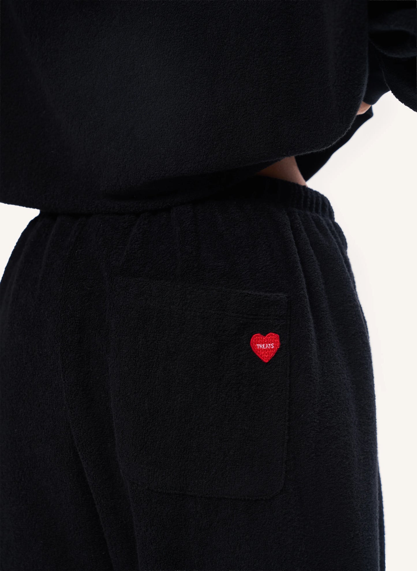 Close-up of a person wearing black fleece sweatpants with an elastic waistband and a side pocket featuring a small red heart logo with white text.