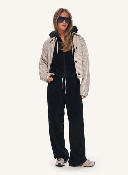A woman modeling a black velour lounge set consisting of a black hoodie and matching wide-leg sweatpants, styled with a beige and black vertically striped zip-up jacket, black sunglasses, and white sneakers against a light off-white background.