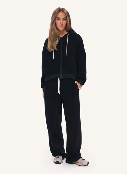A woman modeling a black zip-up hoodie and matching black sweatpants with white drawstrings, standing against a plain light background.