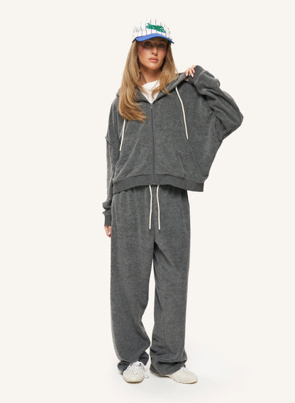 A woman modeling a dark gray zip-up hoodie and matching sweatpants set with white drawstrings, paired with a blue and white baseball cap and white sneakers, against a plain light background.